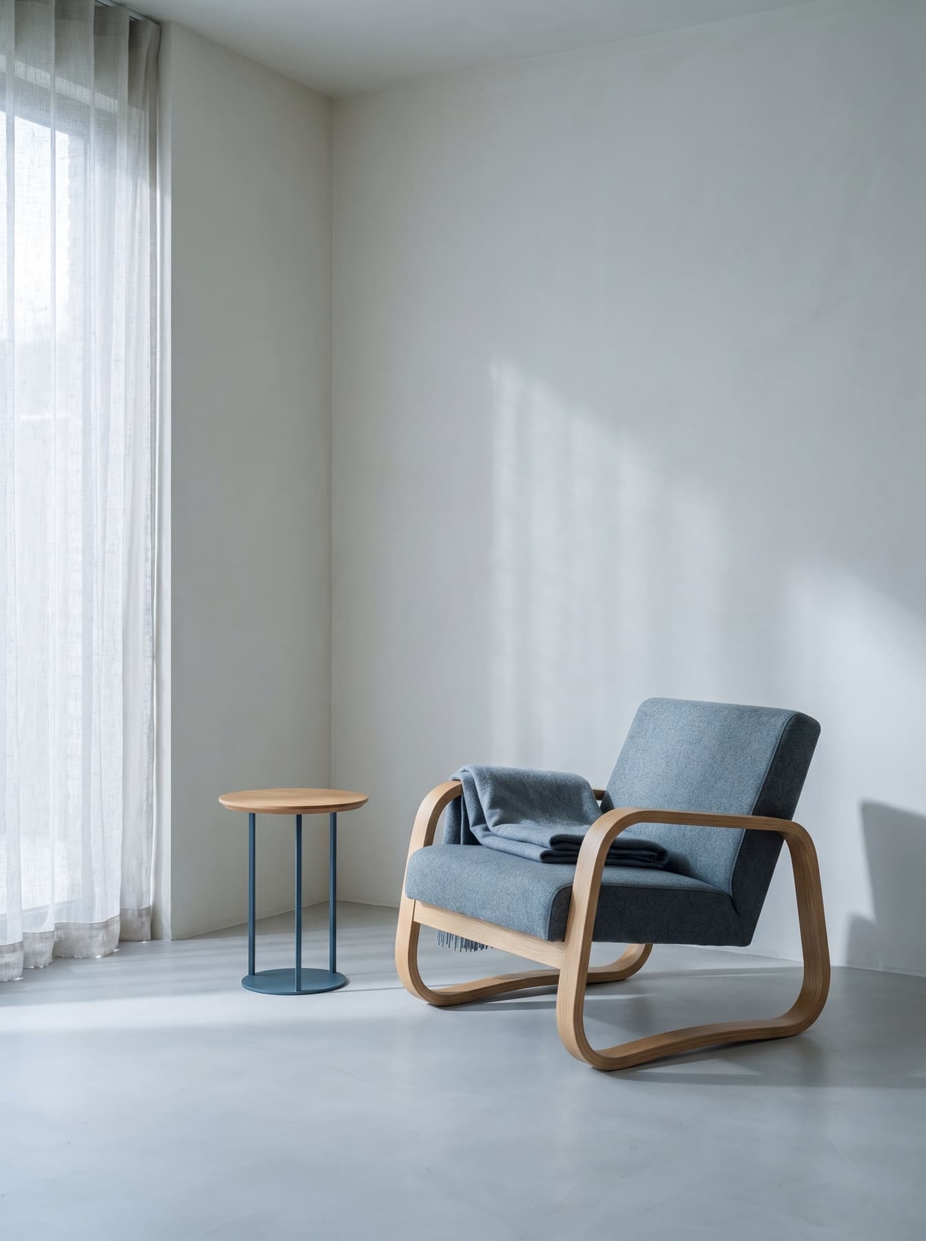 Coin de séjour clair : fauteuil à structure de chêne et assise de laine gris-bleu, guéridon rond à colonne d'acier, rideau de lin devant une fenêtre haute